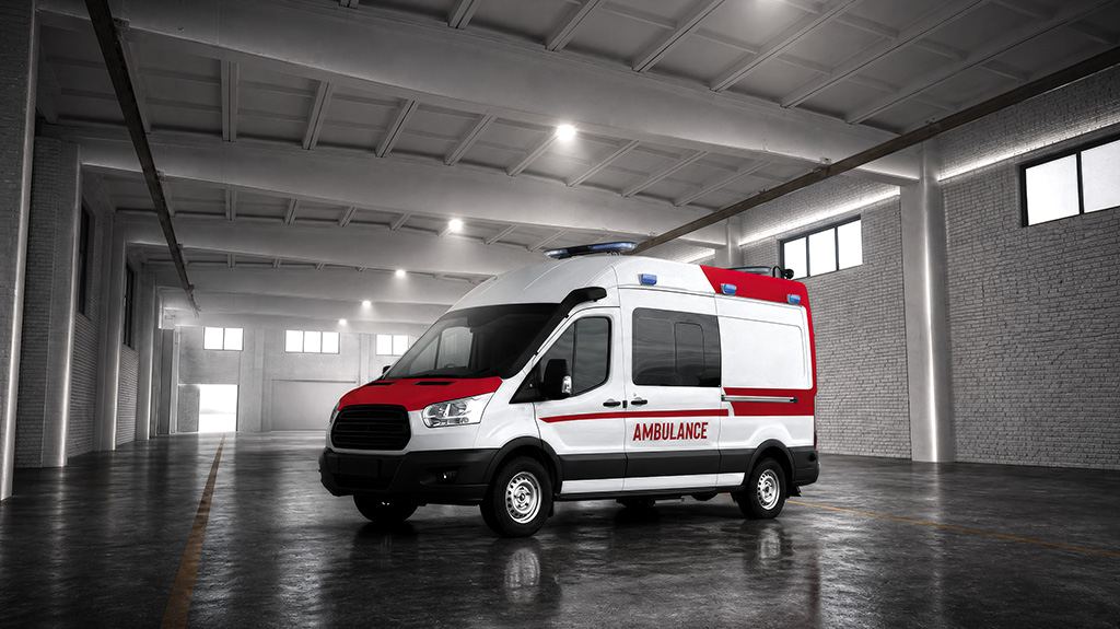An ambulance parked in a large, empty warehouse or garage with a high ceiling and exposed beams, featuring a white body with red stripes and the word "AMBULANCE" written on the side in red letters.