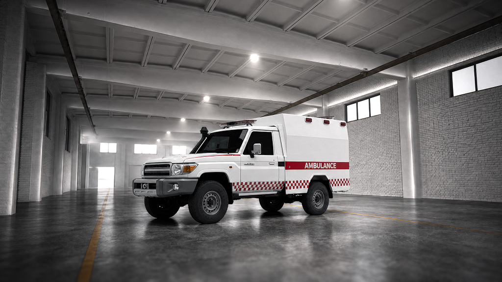 An ambulance in a large warehouse or hangar setting, characterized by a white vehicle with red and white checkered stripes and the word "AMBULANCE" written in red on its side.