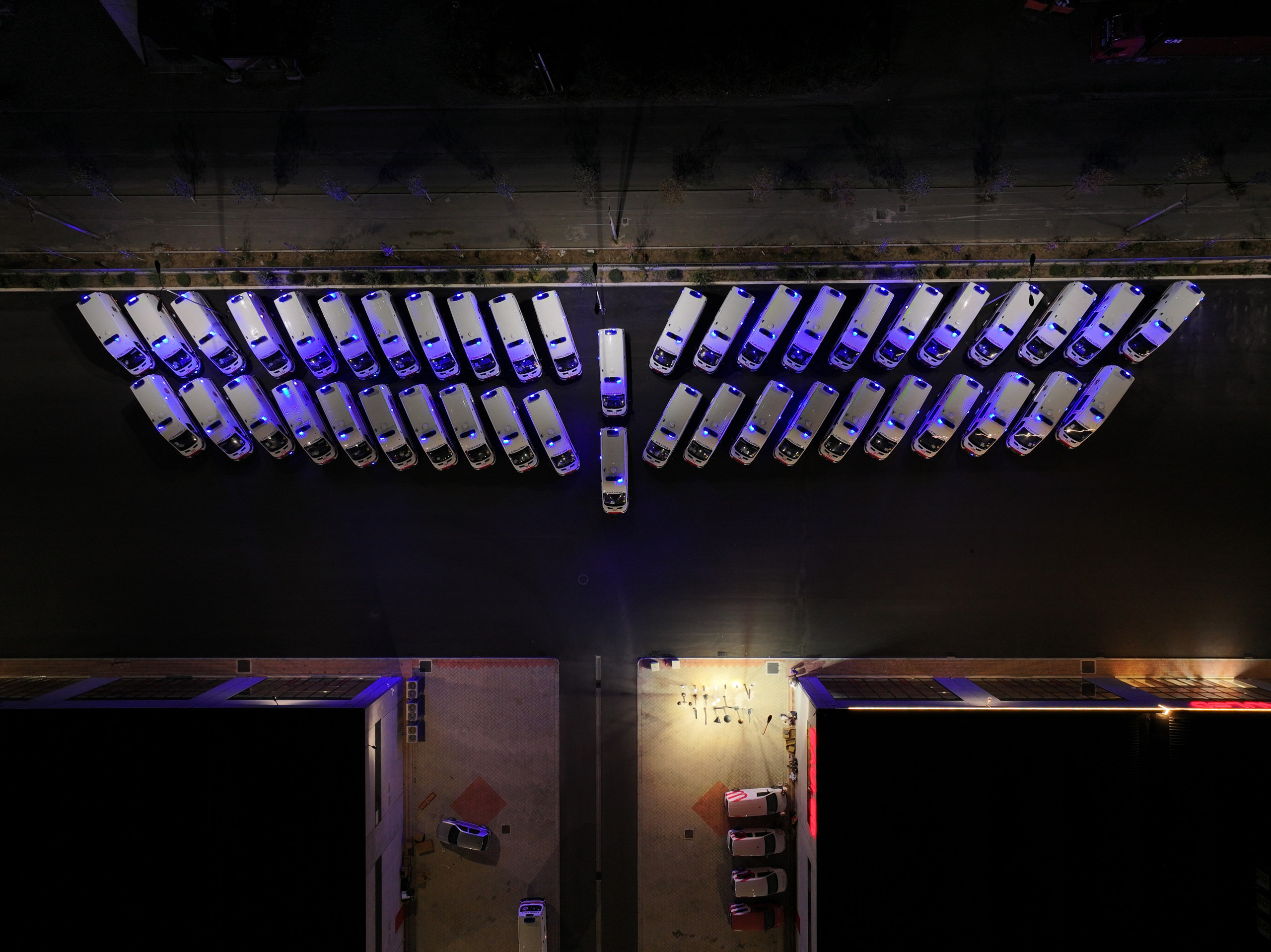 A garage with a row of parked cars, featuring white sedans with blue lights illuminating their roofs, situated on a concrete floor. The garage is illuminated by a single overhead light, creating a dramatic atmosphere. The overall mood of the